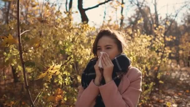 Νεαρή άρρωστη γυναίκα φτερνίζεται στέκεται στο πάρκο την ημέρα του φθινοπώρου, χρησιμοποιώντας μαντήλι - Πλάνα, βίντεο