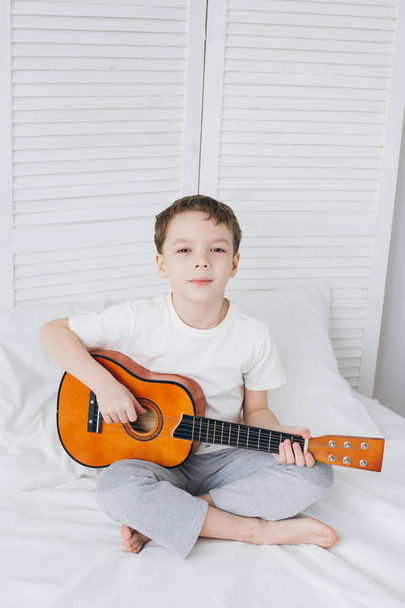 Boy playing a small guitar sitting on the bed  - Photo, image