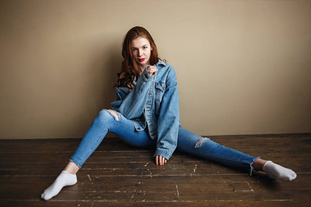 Sensual Female Model Posing in Denim Jacket, Jeans and White Socks on the Floor near Beige Wall. Back to 1990s Fashion Lifestyle Style Concept - Photo, Image