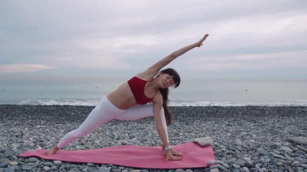 Señora está entrenando el cuerpo por yoga asanas en la orilla del mar en la playa de guijarros durante el día
 - Metraje, vídeo