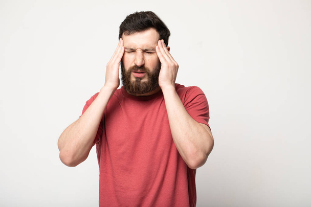 Feeling terrible headache. Portrait of young bearded man is standing and touching his temples while suffering from pain. He is frowning while keeping eyes closed - Photo, image