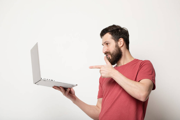 young bearded man looks at laptop screen in his hands and stands isolated on light background - Photo, image