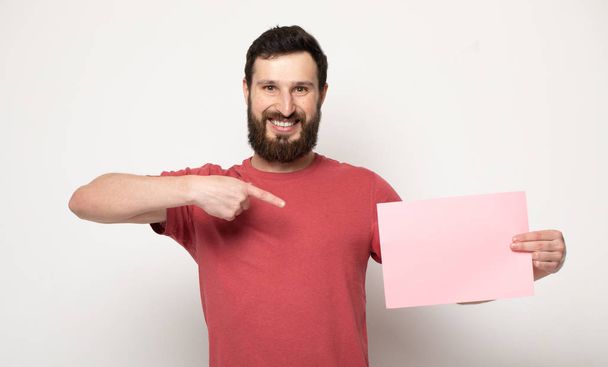 Young bearded man with blank pink banner. Smiling man holding advertising sheet, copy space, isolated - Foto, Imagem