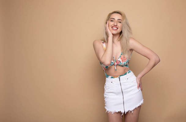 Positive attractive blonde young model wears swimsuit and white skirt being happy to receive good news. Joyful woman rejoices weekends, relaxing indoors, isolated against beige background. - Zdjęcie, obraz