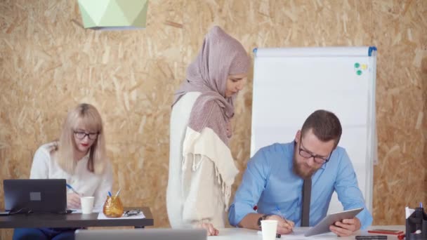 Μουσουλμανική γυναίκα στέκεται κοντά στο τραπέζι του συναδέλφου της ο άνθρωπος στο γραφείο και μιλάει - Πλάνα, βίντεο