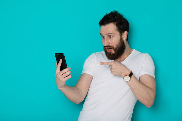 Portrait of a surprised casual man looking at mobile phone isolated over blue background - Φωτογραφία, εικόνα