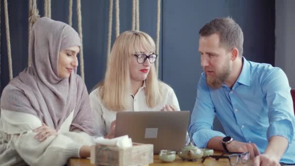 Αραβική γυναίκα και καυκάσιο ζεύγος μιλούν και βλέποντας σε φορητό υπολογιστή στο καφέ - Πλάνα, βίντεο