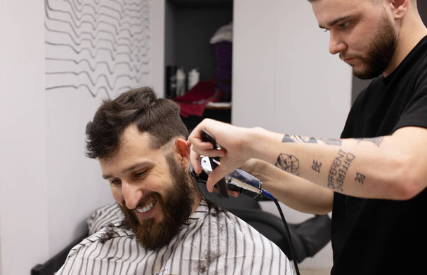 Stylish hairdresser cutting hair of client at barber shop. Beard man getting haircut at salon. - Zdjęcie, obraz