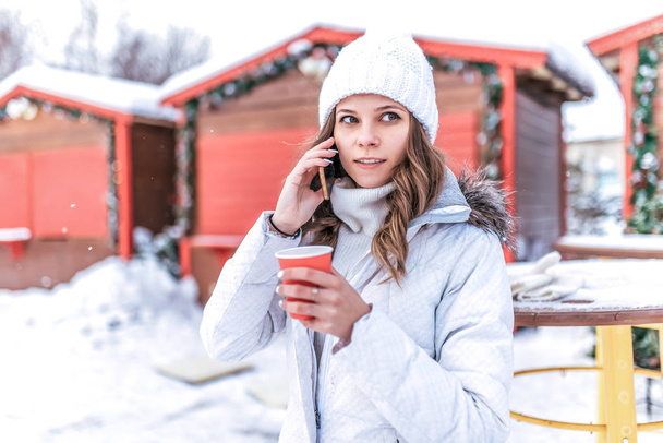 A serious girl in a white jacket and warm hat, talking on phone in the winter in the city in the fresh air. In his hand holds a glass of hot breakfast drink. - Fotó, kép