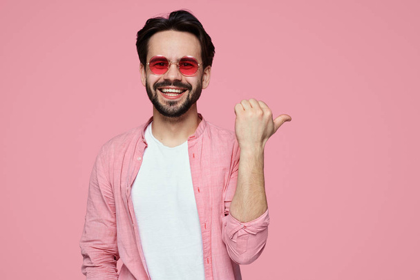 Hndsome giovane ragazzo barbuto in camicia elegante a spazio copia sulla parete rosa come mostra qualcosa di piacevole, ha sguardo sorridente
. - Foto, immagini