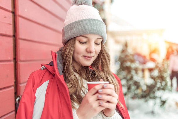 Girl with long hair in winter in city in fresh air. Warms about cup of hot drink coffee tea. In red jacket and warm hat. Emotions of tenderness of comfort pleasant time spending weekend on courtyard. - Фото, зображення