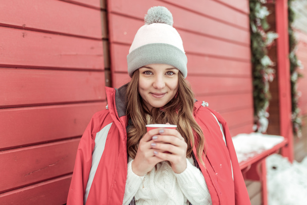 幸せな女が笑みを浮かべて、長い髪冬市の新鮮な空気を持つ少女。熱いカップの温めは、コーヒー紅茶を飲みます。赤いジャケットの温かみのある帽子。中庭の週末感情優しさ快適快適な時間を過ごす. - 写真・画像
