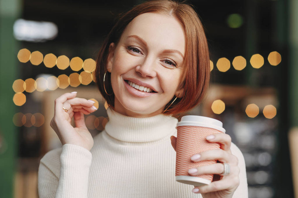 Brown haired woman with make up, toothy smile, dressed in white turtle neck sweater, enjoys hot drink, smiles positively, poses in coffee shop, has make up, expresses happiness. Drinking concept - Photo, Image