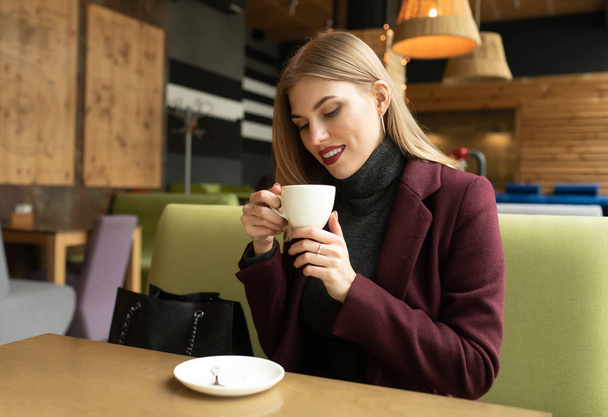 Mooie lachende vrouw die koffie drinkt in het café. Portret van blonde vrouw in een cafetaria die hete cappuccino drinkt en naar de camera kijkt. Mooie vrouw met kopje koffie. - Foto, afbeelding