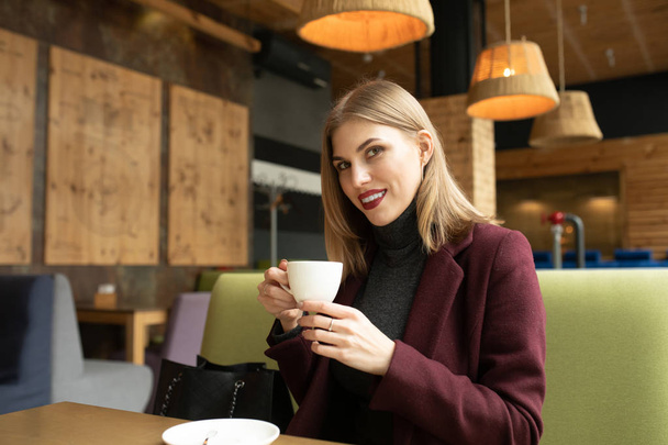 Beautiful smiling woman drinking coffee at cafe. Portrait of blonde woman in a cafeteria drinking hot cappuccino and looking at camera. Pretty woman with cup of coffee. - Foto, afbeelding