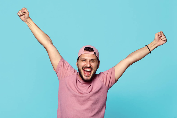 Cheerful young man holding hands up, looks excited, feels triumph, screams with happiness, isolated over pink backdground. - Photo, Image