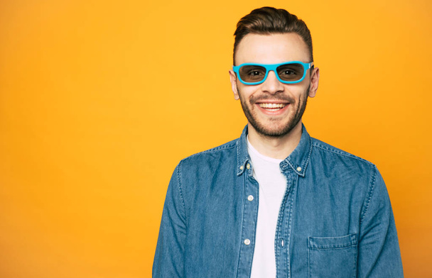 Movie night.  Orange background with a happy man in front of it with a smile and dark brown hair  in denim shirt ,white t-shirt under it  and 3D glasses. - Фото, изображение