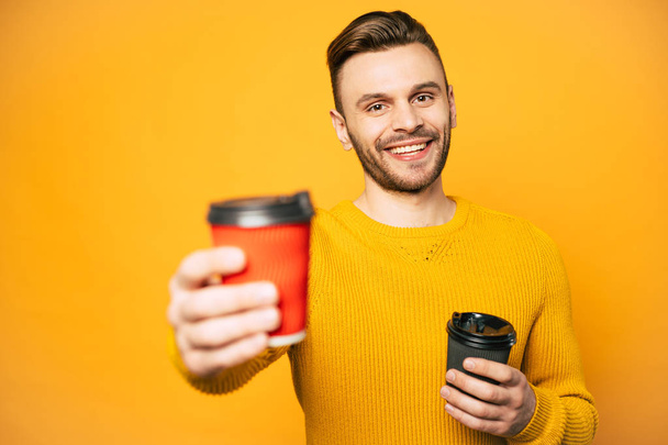 Two cups of coffee or tea in cups of red and black colors in hands of nice-looking man clothed in yellow sweater in front of matching wall. - Fotó, kép