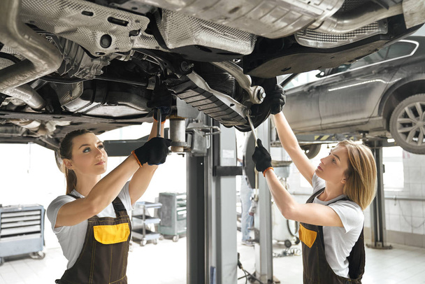 Two girls fixing lifted auto undercarriage, using wrenches. - Valokuva, kuva