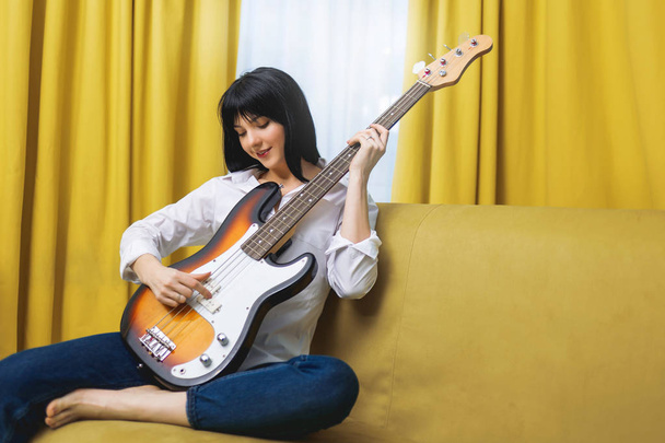 Pretty young woman having her legs crossed playing some record or exercise on bass guitar sitting on couch at home lifestyle - Photo, Image