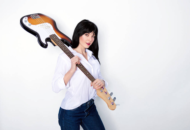Atraktivní brunetka držící svou basovou kytaru na rameni přes šedé pozadí - Fotografie, Obrázek