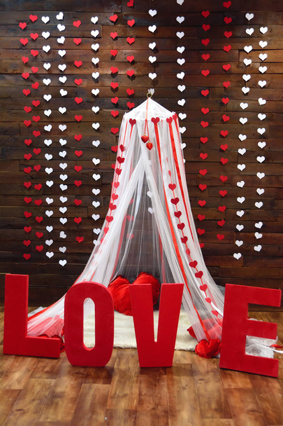 Wooden wall. Paper garlands in the shape of hearts. White canopy. Red heart pillows. Decorative white carpet. Romantic photo zone. Red three-dimensional letters of foam. - Photo, Image