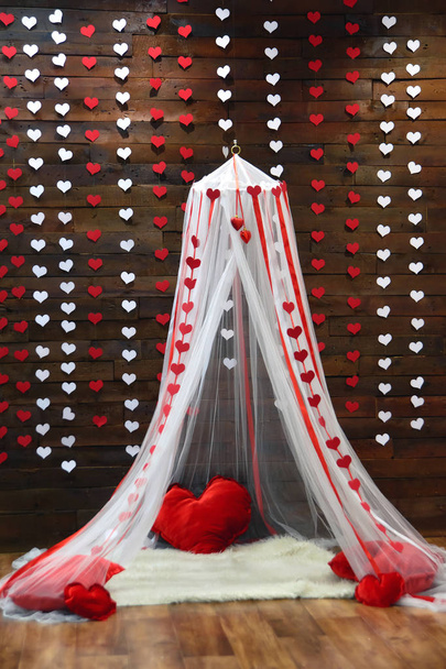Wooden wall. Paper garlands in the shape of hearts. White canopy. Red heart pillows. Decorative white carpet. Brown linoleum. Romantic photo zone. - Photo, Image