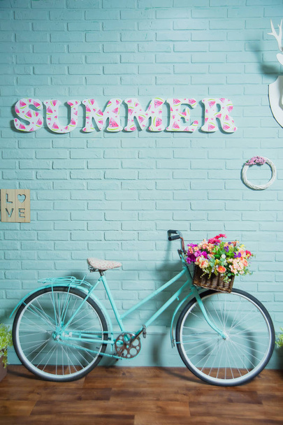 Ποδήλατο με ένα καλάθι λουλουδιών. Μπλε τοίχος από τούβλα. Καλοκαιρινή διακόσμηση. Κορνίζες για φωτογραφίες στον τοίχο. Λινέλαιο με τη μορφή παρκέ. Καλάθια πράσινων φυτών. Διακοσμητικά τούβλα. - Φωτογραφία, εικόνα