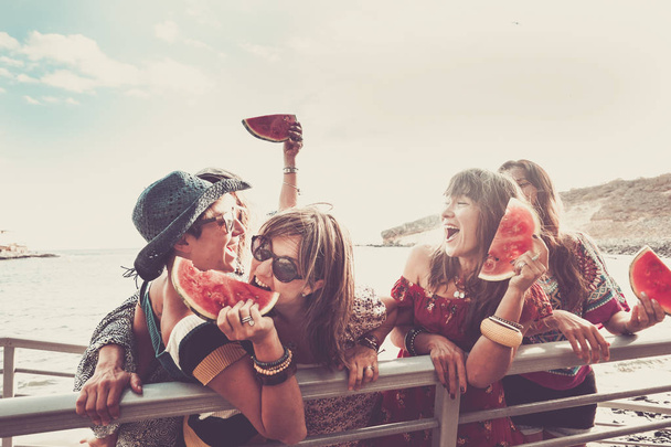 Grupa młodych wesołych dziewcząt bawi się razem jedząc razem arbuza na plaży z oceanem i czystym niebem w tle - przyjaźń i zabawni ludzie w wolnym czasie na świeżym powietrzu - Zdjęcie, obraz