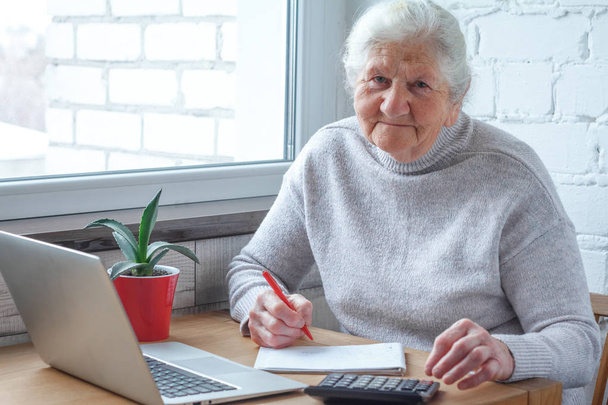 Eine alte Frau sitzt am Tisch vor dem Laptop. - Foto, Bild