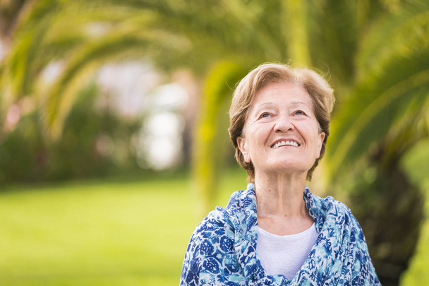 Retrato de la alegre y cauta mujer mayor caucásica sonriendo y mirando hacia arriba - al aire libre naturaleza verde fondo tropical - camisa azul y pelo rubio. - Foto, imagen