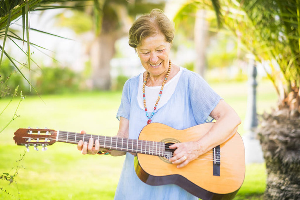 Cheerful and happy old aged senior caucasian woman smiling and having fun playing a  guitar and dong music in outdoor park leisure activity - Photo, Image
