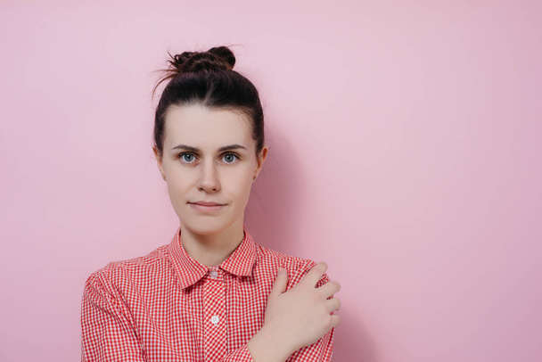 Positive shy young girlfriend has tender smile, keeps hand on shoulder and energized at camera feeling optimistic gazing trustworthy at camera over pink background - Photo, Image