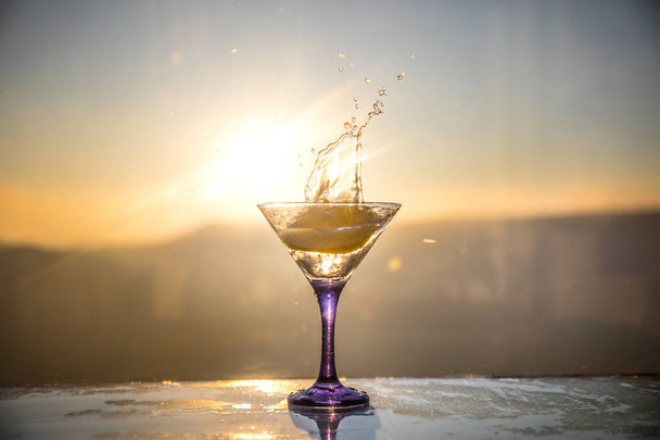 Copa de cóctel Martini salpicando sobre fondo nebuloso al atardecer o cóctel colorido en vaso con salpicaduras y limón. Entretenimiento al aire libre al aire libre. Enfoque selectivo
 - Foto, imagen