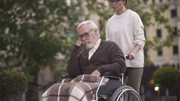 Young woman on walk with sad pensioner in wheelchair, caregiver assistance, help - Foto, Imagem