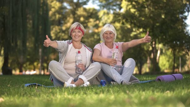 Dos mujeres mostrando los pulgares hacia arriba sentado en la hierba después de hacer ejercicios, positivo
 - Foto, imagen