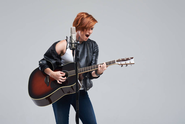 Portrait of a young woman musician with an acoustic guitar. Sing a song on studio microphone on a gray background. She wears black leather jacket and plays rock and roll loudly. Space for text - Photo, Image