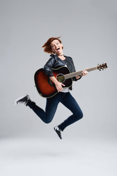 Young woman musician with an acoustic guitar in hand on a gray background. She laughs and jumps high. plays rock and roll loudly. Full-length portrait.  - Fotoğraf, Görsel