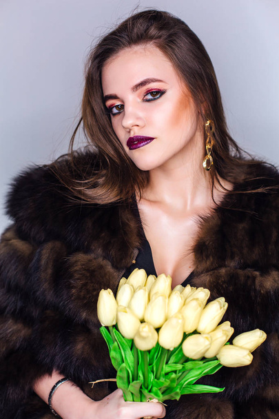 Beauty portrait of a woman with bright colorful make-up dressed in a fur coat holding bouquet of yellow tulips - Foto, afbeelding