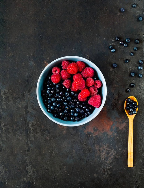 Bilberry and raspberries in blue bowl on vintage rusty metal background. Concept of organic berries.  - Photo, image