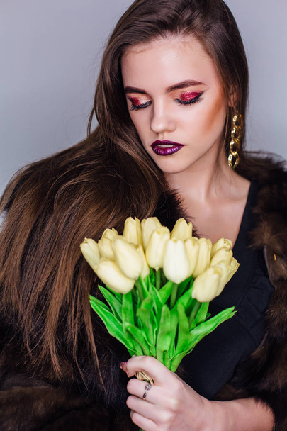 黄色のチューリップの花束を持つ毛皮のコートに身を包んだ明るいカラフルなメイクアップを持つ女性の美しさの肖像画 - 写真・画像