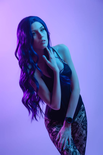 Hermosa mujer con el pelo largo de color púrpura en ropa interior negra, pantimedias posando en posición atractiva en coloridas luces de color azul neón y rosa en el estudio. Concepto de moda
 - Foto, imagen