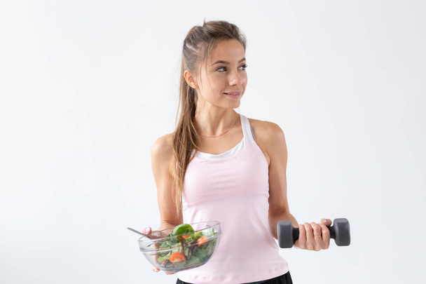 Healthy lifestyle, people concept - a plate of salad in one hand and dumbbell in another hand. Smile and fitness suit - Photo, image