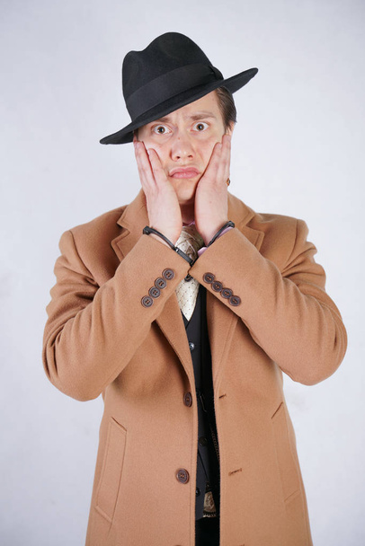 молодой человек в черном деловом костюме и бежевом модном пальто в стильной шляпе стоит в наручниках на белом фоне студии
 - Фото, изображение