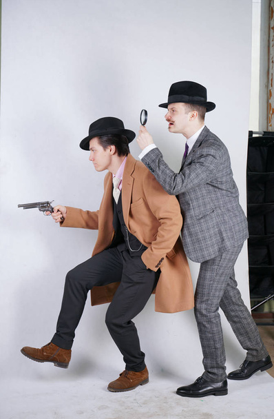 two caucasian detective men in retro suits with vintage hats sneak quietly go, holding a magnifying glass and a gun on white studio background - Fotografie, Obrázek