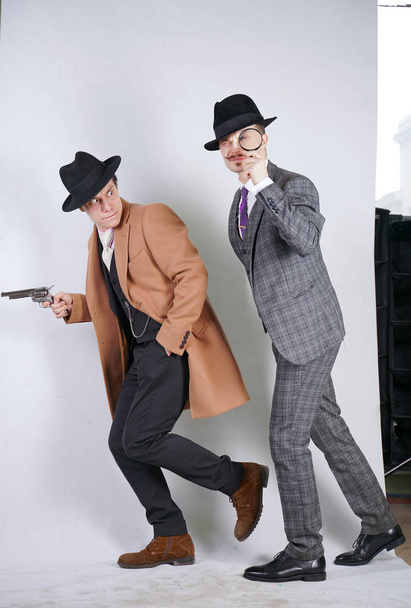 two caucasian detective men in retro suits with vintage hats sneak quietly go, holding a magnifying glass and a gun on white studio background - Photo, Image