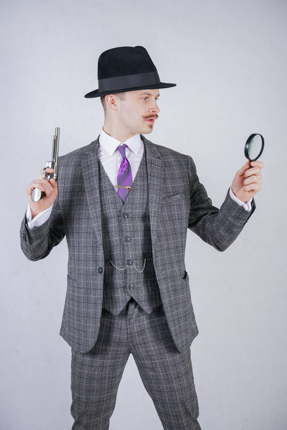 man with a mustache dressed in a business plaid suit with a tie, holding a magnifying glass and a gun on a white solid Studio background - Photo, Image
