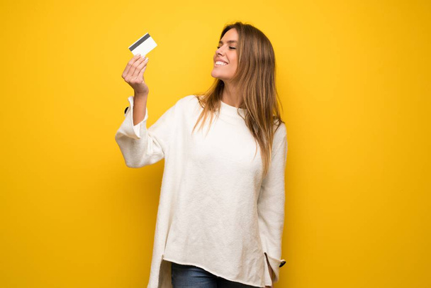 Mujer rubia sobre pared amarilla sosteniendo una tarjeta de crédito y pensando
 - Foto, Imagen
