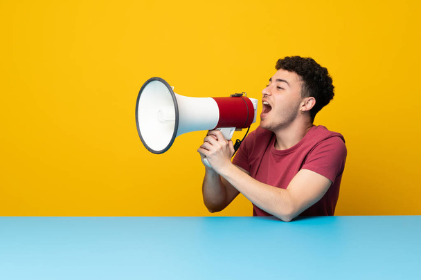 Jonge man met kleurrijke wand en tafel schreeuwen door een megafoon - Foto, afbeelding