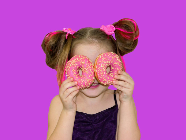 A little kid girl dabbles and plays with two fresh donuts before eating. A child holds donuts near his eyes and looks through the holes like through glasses. - Foto, imagen